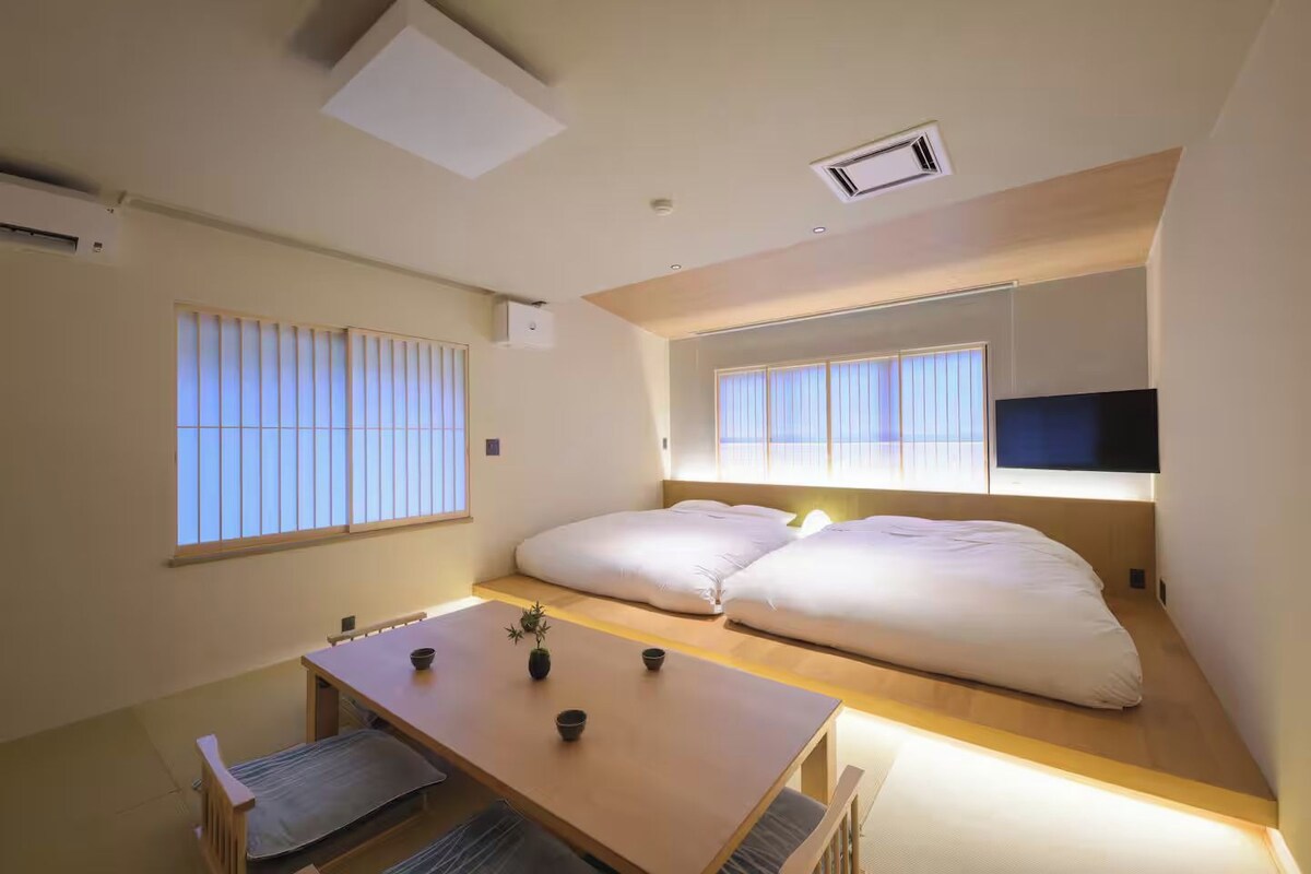 Quarto de hotel pequeno, com cama, televisão, mesa e janela com persianas.