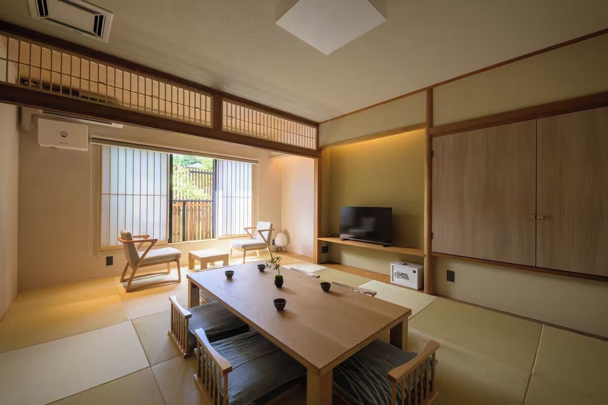 Quarto japonês tradicional com tatames, uma mesa baixa com almofadas, uma televisão em uma estante e portas deslizantes.