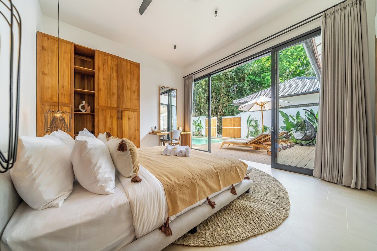 A bedroom with a large bed, wooden wardrobe, and a view of a pool area.
