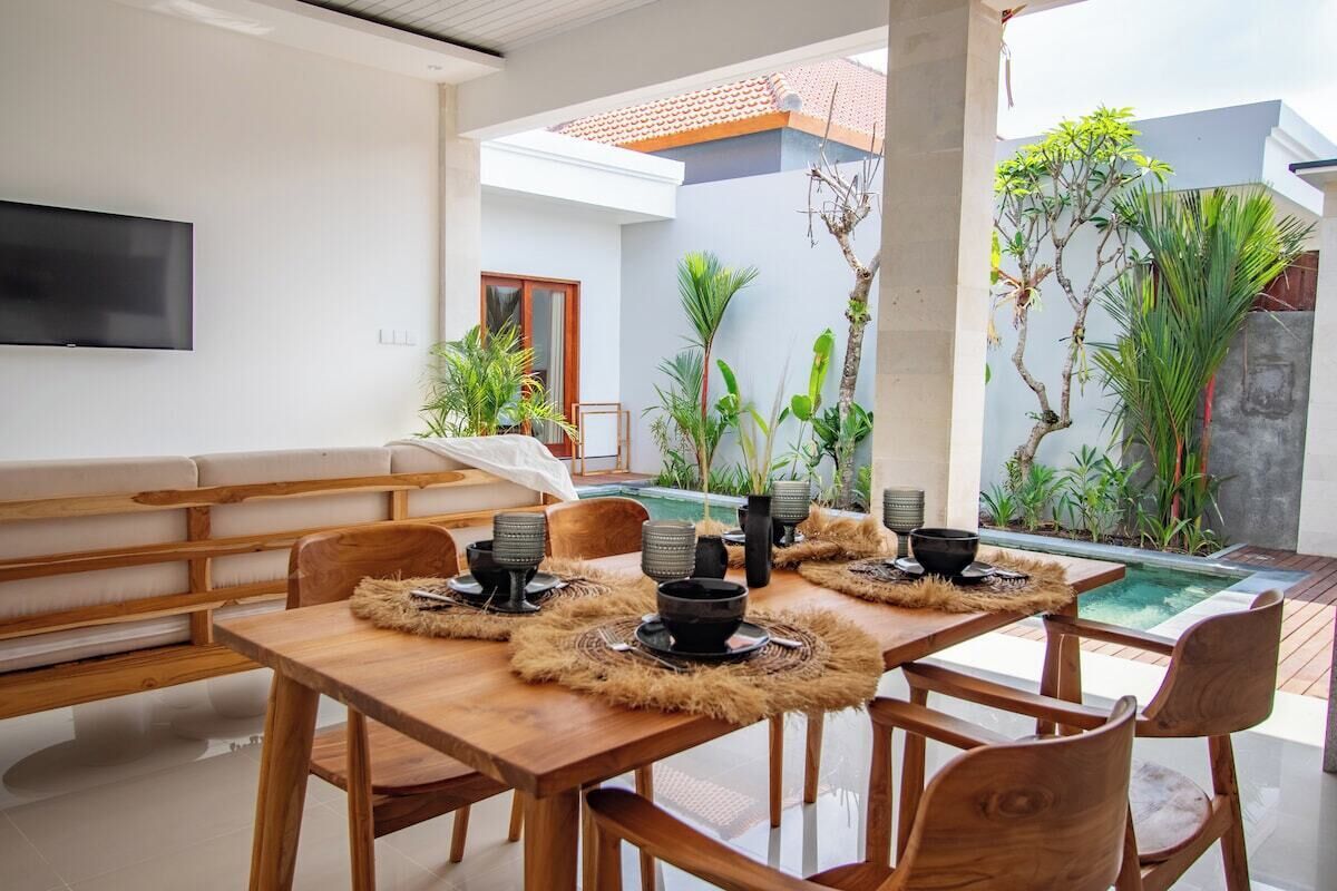 A dining area with a wooden table and chairs, a TV mounted on the wall, and a pool in the background.