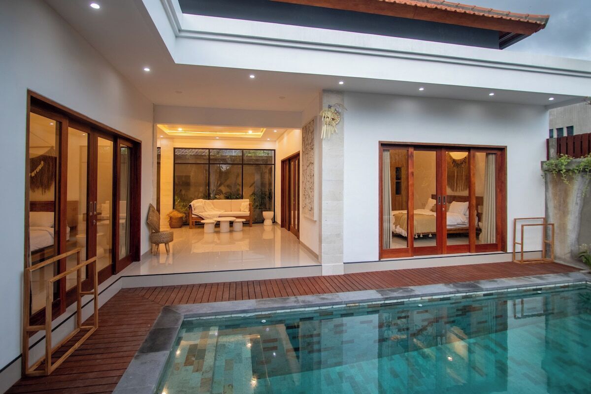 A modern indoor swimming pool area with a glass enclosure, wooden deck, and a cozy seating area with cushions.