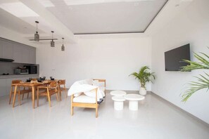 A modern living room with a sofa, dining table, and chairs, a kitchen area, and a mounted TV on the wall. - Cozy 2BR Paradise w Beach Nearby & Huge Pool+Patio (Kabupaten Badung)