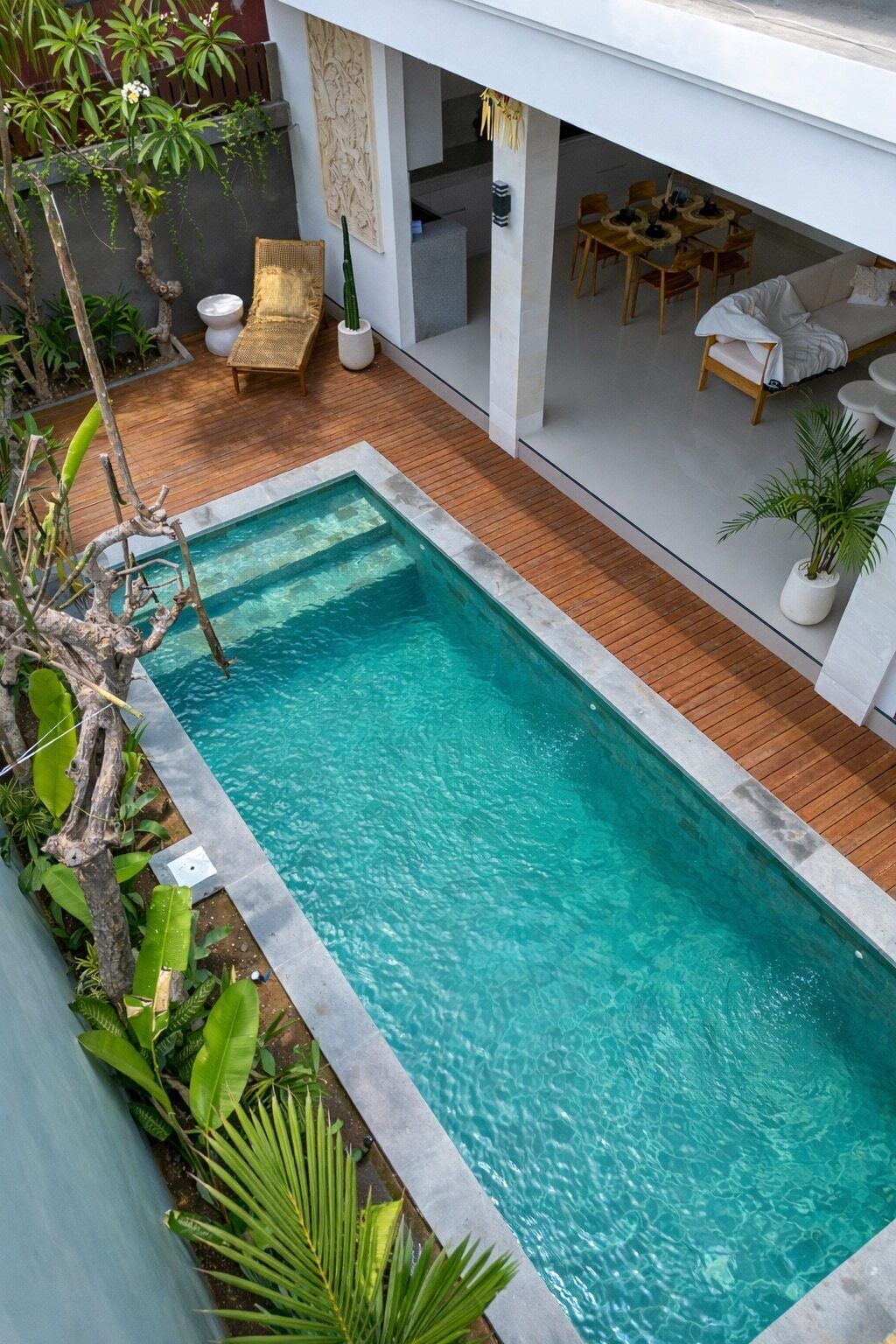 Piscina retangular com deck de madeira ao redor, área de estar com cadeiras e mesa, e parede branca com design texturizado.