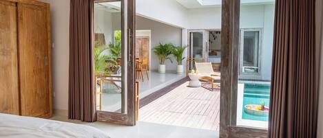 A bedroom with a large bed, a wooden wardrobe, and a sliding glass door leading to a balcony with a pool.