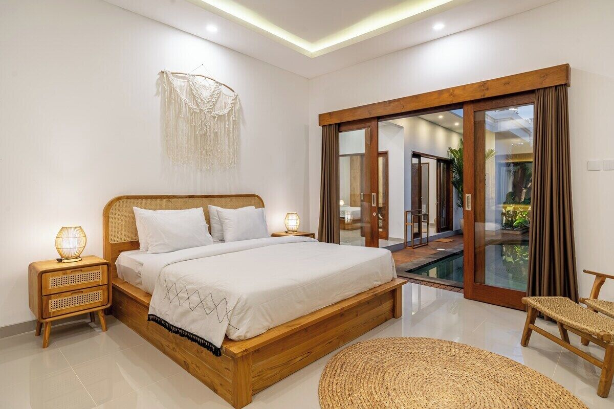 Quarto moderno com cama de madeira, porta de vidro para uma varanda e uma cadeira de fibra natural.