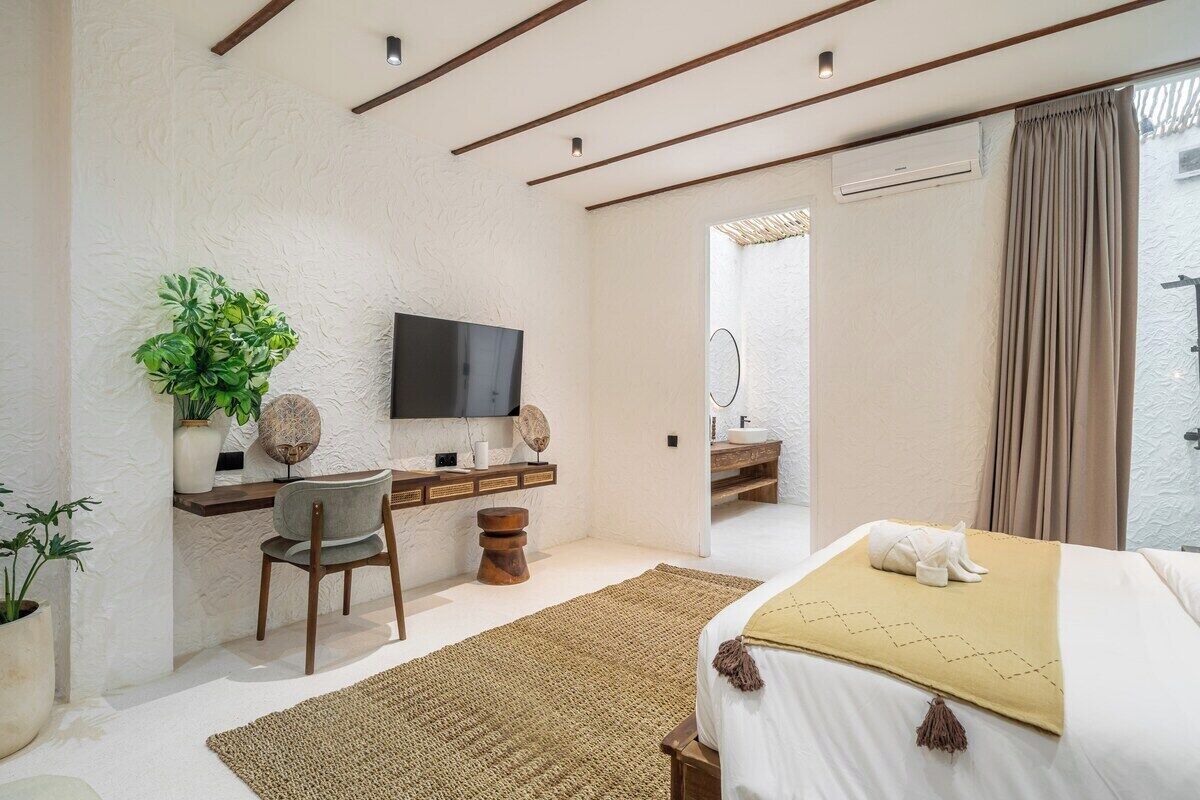 Ein Schlafzimmer mit Bett, Schreibtisch, Stuhl, Fernseher und einem durchgegangenen Tür sichtbar liegenden Badezimmer.