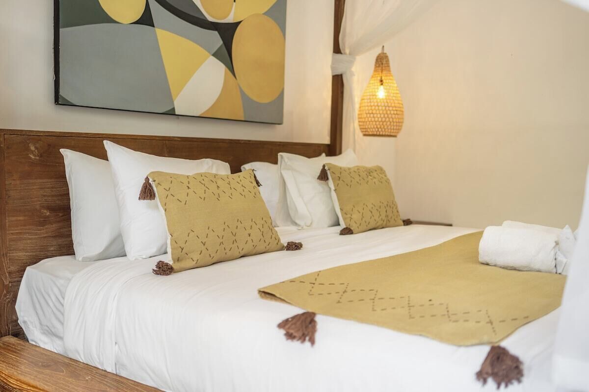 Un lit bien fait, avec du linge de lit blanc et des coussins décoratifs, un tableau abstrait accroché au mur et un luminaire suspendu de style lanterne.