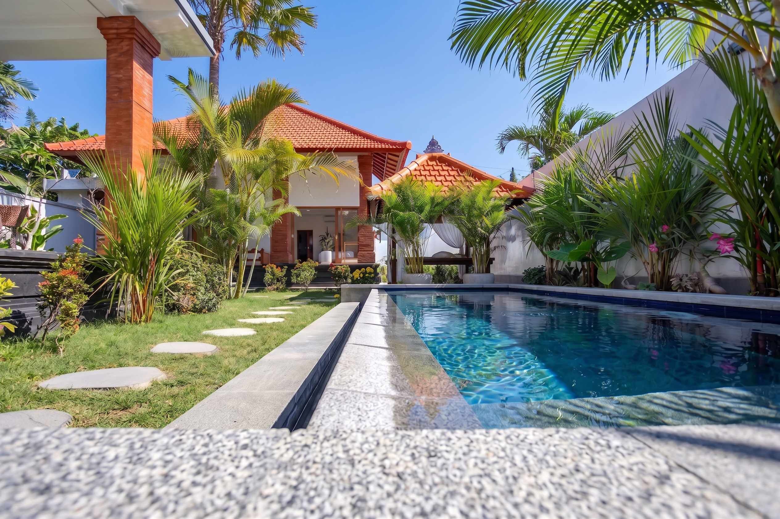 Une piscine à l’eau d’un bleu limpide, entourée d’un jardin bien entretenu avec des palmiers et un sentier menant à un bâtiment dont le toit est recouvert de tuiles rouges.
