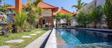 A swimming pool with a clear blue water surface, surrounded by a well-maintained garden with palm trees and a pathway leading to a building with a red-tiled roof.