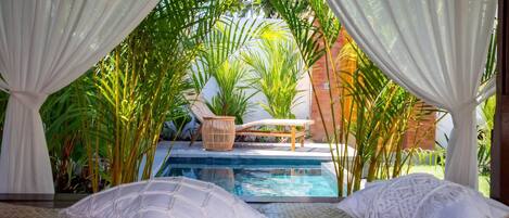 A private pool area with a lounge chair and a small table, surrounded by lush greenery and white drapes.