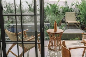 Blick durch eine Glastür in einen Raum mit Rattanmöbeln, einem runden Tisch und einem Pool im Freien.
