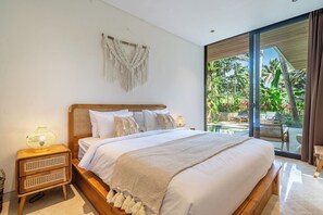 Ein Schlafzimmer mit einem großen Bett, einem Nachttisch, einer Wanddeko aus Makramee und Blick auf einen tropischen Garten durch eine Glastür.