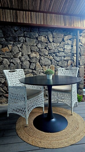 Een ronde tafel met een zwart onderstel, omringd door witte rieten stoelen, voor een achtergrond van een stenen muur.