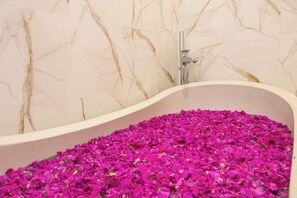 Eine Badewanne, gefüllt mit rosa Blütenblättern, eine Marmorwand und ein moderner Duschkopf.
