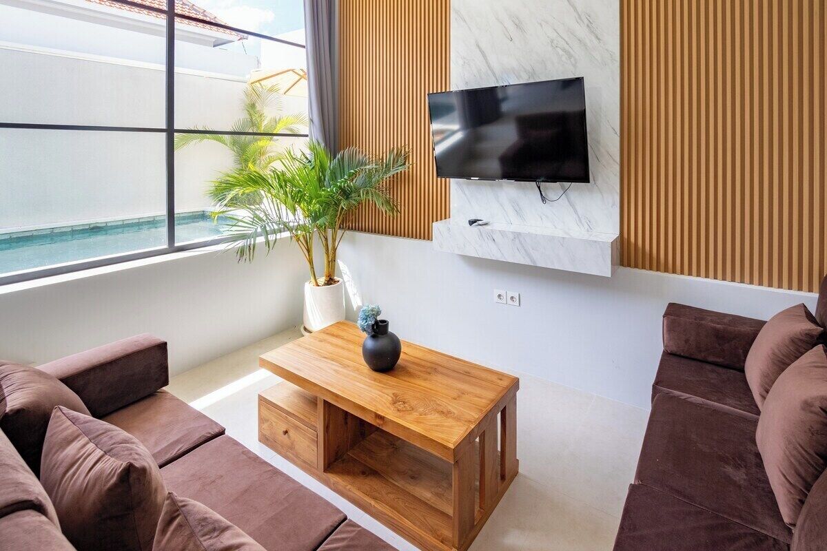 Uma sala de estar moderna com sofá, mesa de centro de madeira, planta em vaso e televisão na parede.