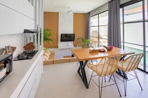 Une cuisine moderne dotée d’une cuisinière intégrée, un coin repas avec une table en bois et des chaises en osier, un téléviseur à écran plat fixé au mur et une grande fenêtre avec des rideaux.
