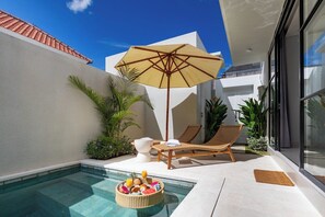 Ein moderner Poolbereich im Freien mit einer Liege, einem Korbwicker-Korb mit Früchten und einer Flasche sowie einem Sonnenschirm.