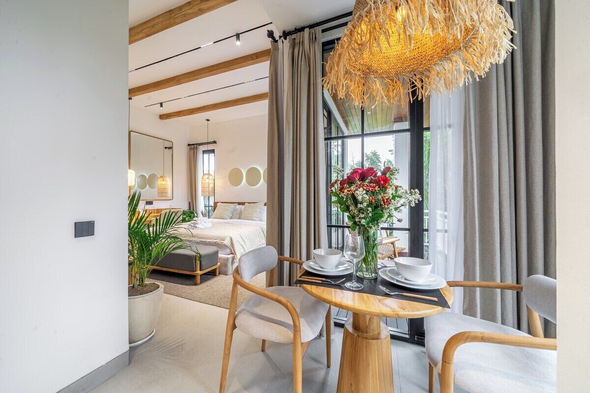 Una habitación de hotel moderna con zona de comedor, un ventanal con cortinas y una lámpara colgante.
