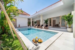 Una piscina amb gandules, taula de dinar amb fruita i begudes, i un jardí tropical.