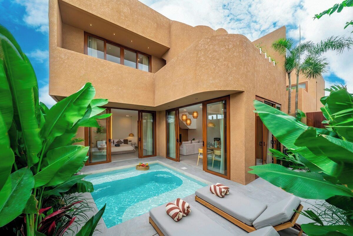 A modern building with a swimming pool, lounge area, and glass doors leading to an interior space.