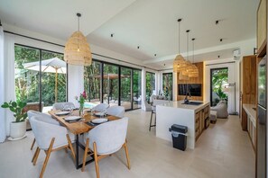 A modern kitchen with a central island, large windows, and a dining area with a table and chairs. - Ubud 2BR Escape w/Pool! Caffes & Nature Nearby (Gianyar Regency)