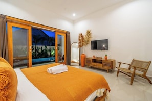 A hotel room with a bed, a TV, a wooden dresser, a wicker chair, and a sliding glass door leading to a balcony. - Brand new stylish villa, Huge pool, Near the beach (Kecamatan Kuta Utara)
