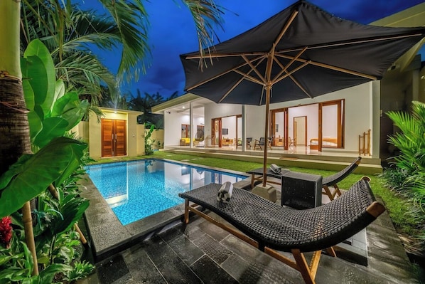 A swimming pool with lounge chairs and an umbrella, surrounded by tropical plants. - Brand new stylish villa, Huge pool, Near the beach (Kecamatan Kuta Utara)
