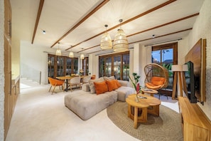 A spacious living area with a large sofa, a dining table, and a TV. There are wicker chairs and a hanging chair. The room has wooden beams on the ceiling and large windows. - Brand New Oasis Villa, Large Pool, Central Ubud (Kabupaten Gianyar)