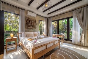 Ein geräumiges Schlafzimmer mit einem großen Bett, einem Holz-Kopfteil, einer Wandleuchte und Blick auf Grün hinter großen Fenstern.