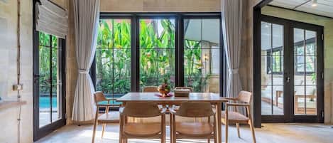A dining area with a table and chairs, large windows with curtains, and a view of greenery outside.