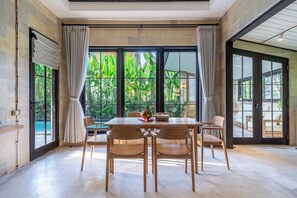 A dining area with a table and chairs, large windows with curtains, and a view of greenery outside. - Cozy Ubud's 2BR Escape with Huge Pool & Patio (Ubud)