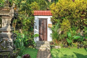 Un pequeño edificio con techo rojo y una puerta, rodeado de exuberante vegetación y una estructura de piedra a la izquierda.
