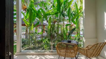 Une pièce dotée d’une grande fenêtre donnant sur un jardin tropical, de deux fauteuils en osier et d’une petite table.