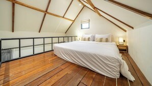 Quarto com teto inclinado, vigas de madeira, cama, mesa de cabeceira e janela.
