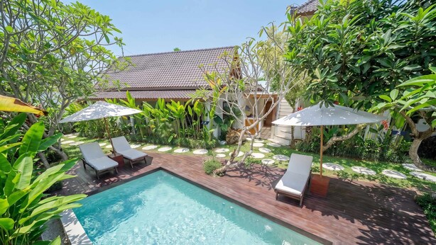 Piscina retangular com duas espreguiçadeiras e guarda-sóis, cercada por vegetação exuberante e um edifício com telhado de azulejos.