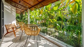 Uma área de estar coberta ao ar livre, com cadeiras de vime e uma mesa, rodeada por plantas tropicais exuberantes.
