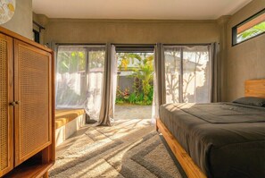 Ein Schlafzimmer mit einem Bett, einem Rattanschrank und Blick auf einen Garten durch eine Schiebetür.