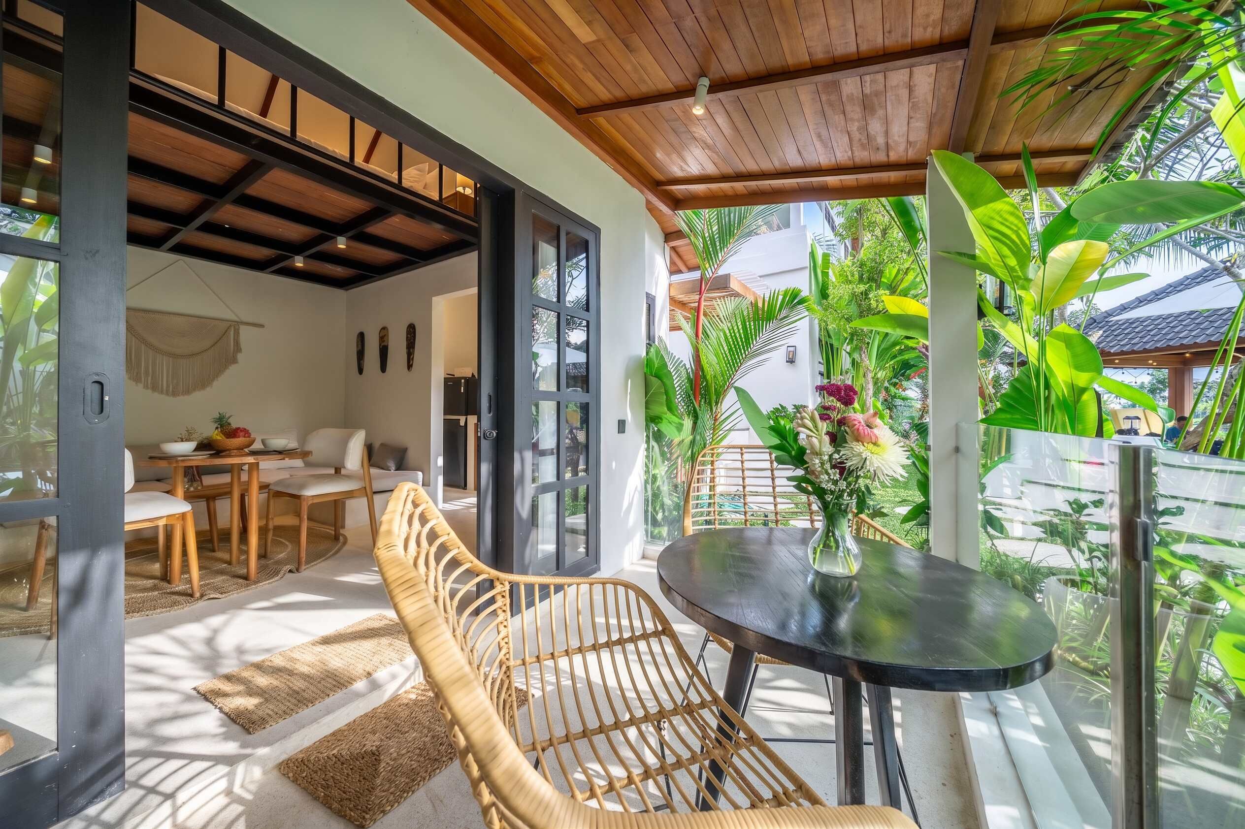 A modern outdoor seating area with a round table and wicker chairs, a dining table with chairs, and a view of lush greenery and a traditional house in the background.