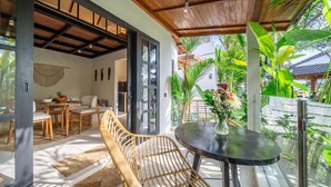 A modern outdoor seating area with a round table and wicker chairs, a dining table with chairs, and a view of lush greenery and a traditional house in the background. - Cozy Ubud 1BR w Open-Air Bath, Pool & Garden (Gianyar Regency)