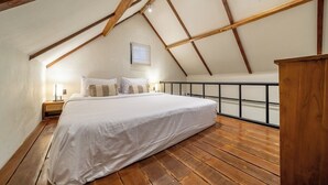 A bedroom with a sloped ceiling, wooden beams, a bed with white bedding, a nightstand, and a window. - Cozy Ubud 1BR w Open-Air Bath, Pool & Garden (Gianyar Regency)