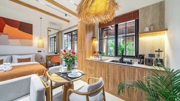 A hotel room with a bed, a dining table with chairs, a kitchen area, and a large window with a view of greenery.