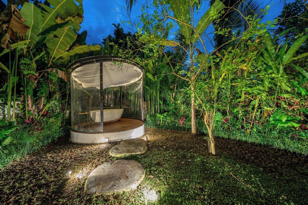 A circular glass structure with a wooden bench inside, surrounded by lush greenery and a clear sky.
