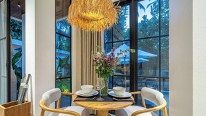 Un comedor con una mesa de madera redonda, dos sillas blancas tapizadas, un florero con flores y una lámpara colgante.