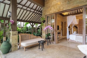 A covered outdoor seating area with a wicker sofa, a small table, and potted orchids. - Cozy 1BR in Lush Ubud w Garden Bath • Pool • Patio (Gianyar Regency)