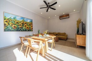 Ein Essbereich mit einem Holztisch und Stühlen, einem Sonnenblumenbild an der Wand, einem Deckenventilator und einer Couch im Hintergrund.