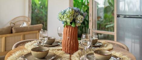 A dining area with a round table set for four, surrounded by woven placemats, and a vase with flowers in the center.