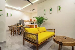 Un salon moderne avec un canapé en osier, une table à manger avec des chaises, un coin cuisine et une grande plante.