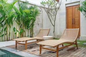 Zwei Korbsessel am Pool mit einer Holzterrasse, umgeben von tropischen Pflanzen und einer weißen Wand mit einer Holztür.