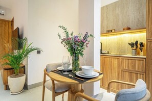 A dining area with a table set for two, a vase with flowers, and a kitchen in the background. - Cozy 1BR • Pool&Garden View • Patio • Central Ubud (Kabupaten Gianyar)