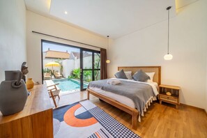 A modern bedroom with a large bed, a wooden nightstand, a desk with a vase, and a view of a swimming pool. - Paradise 2BR Villa w Beach Access & Private Pool (Kecamatan Kuta Utara)